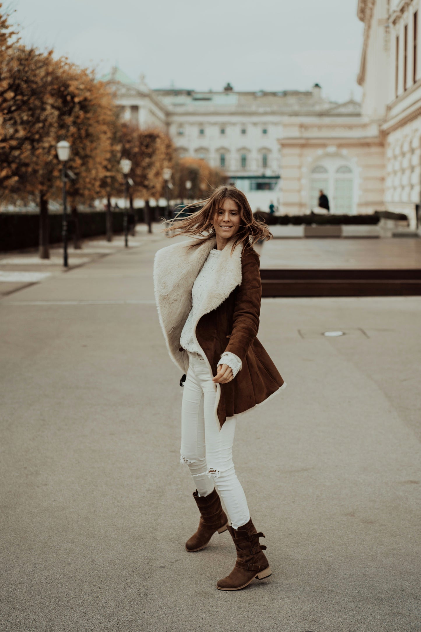 Woman in a brown coat with white fur lining standing in an urban setting.