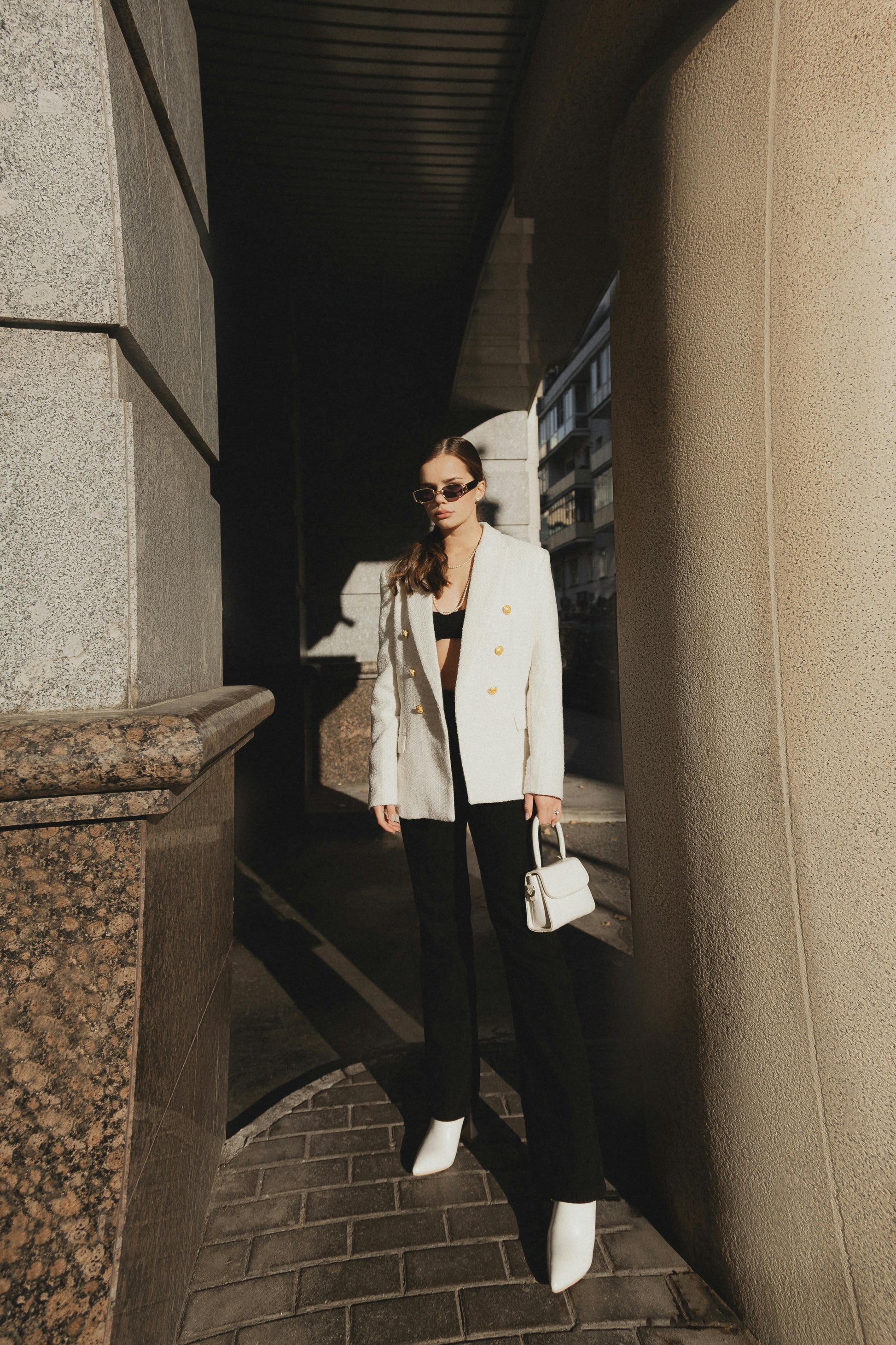 Woman in a white blazer and black pants standing between two buildings on a city street.