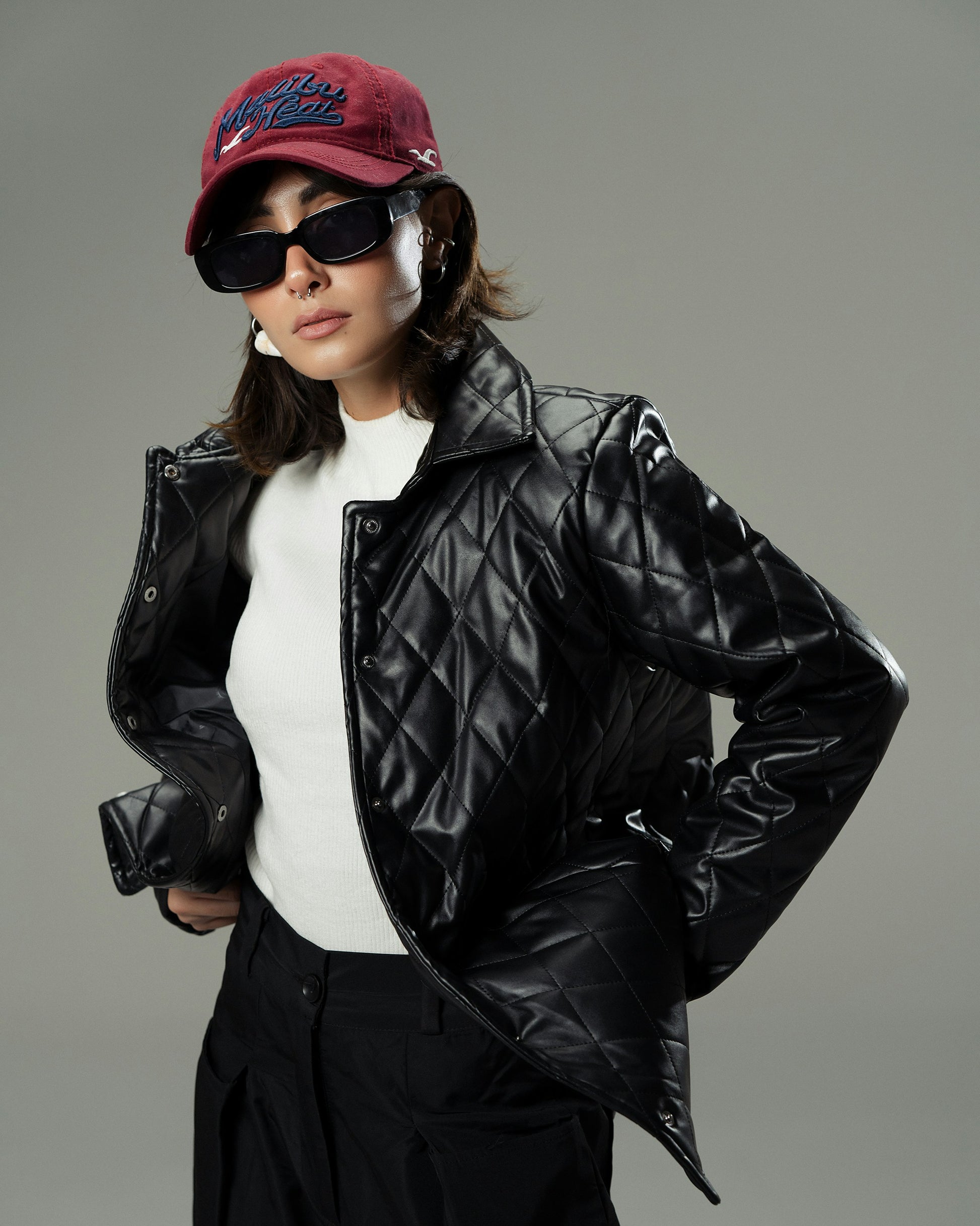 Person wearing a black quilted jacket, white shirt, dark pants, sunglasses, and a red cap on a gray background
