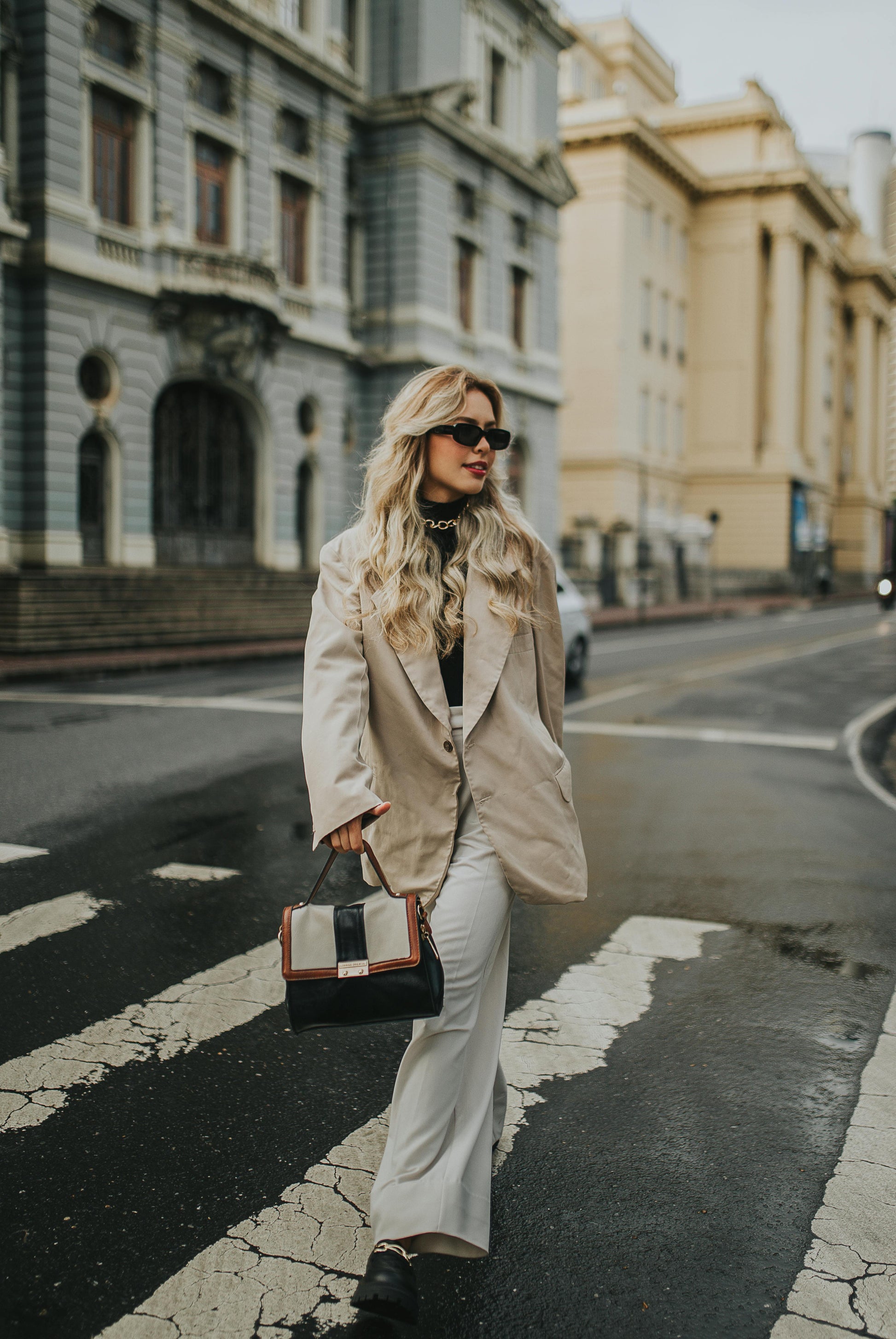 Woman in a beige coat and white pants walking on a city street.