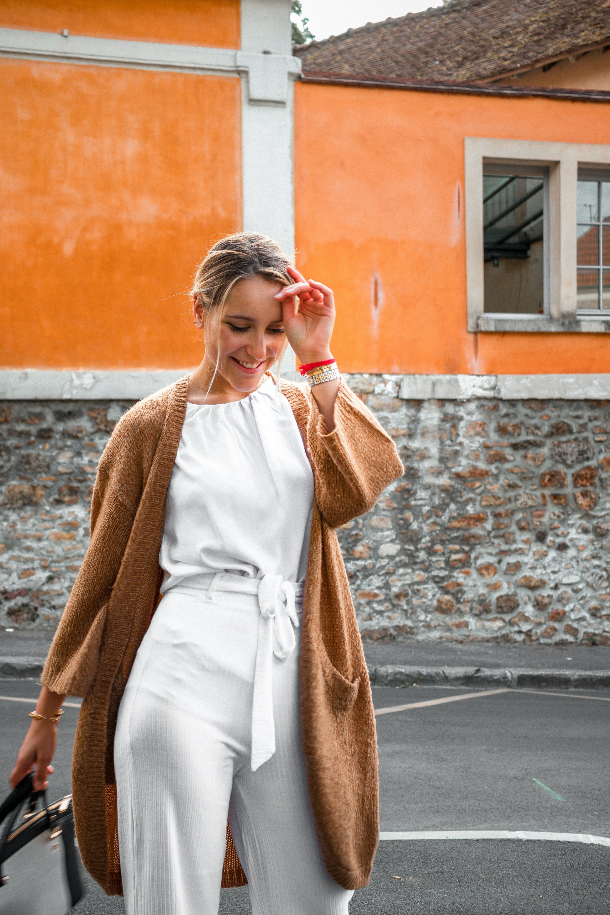 Woman in a brown cardigan and white outfit walking in front of an orange building.