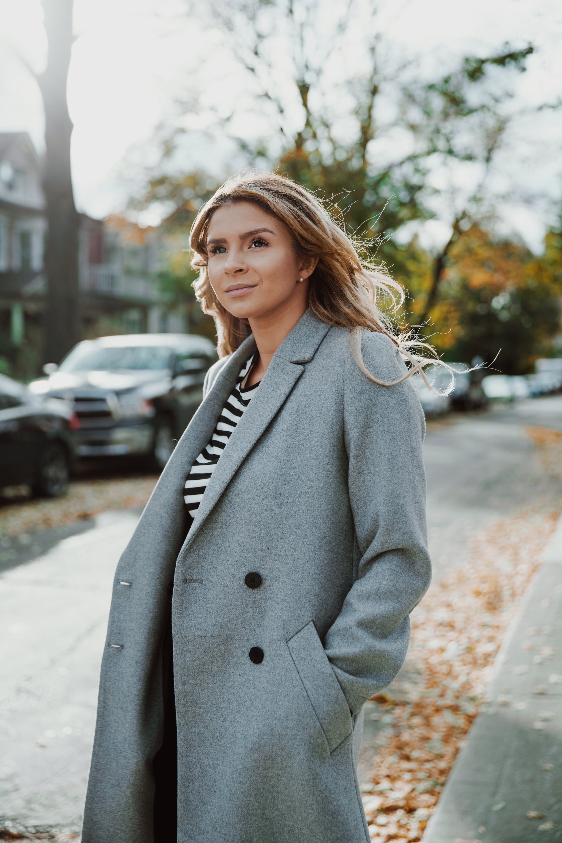 Woman wearing a gray coat standing on a street with trees and cars in the background