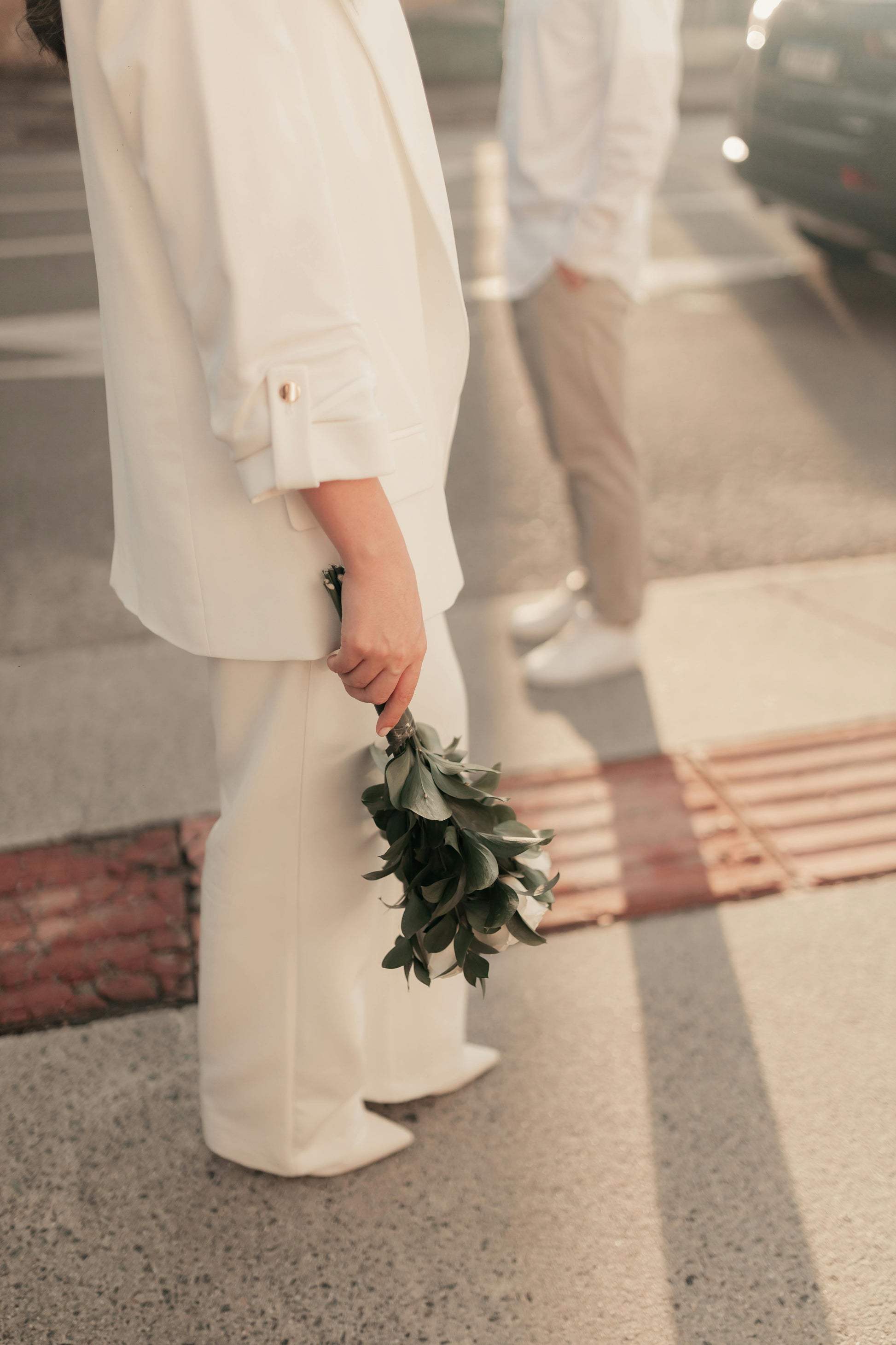 Person in a white outfit holding a small bouquet on a street.