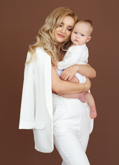 Woman in white holding a baby against a brown background. Victoria Ellison is a Fashion Stylist specialising in Bridal Styling, Pregnancy Styling, Motherhood Styling, Personal Styling.